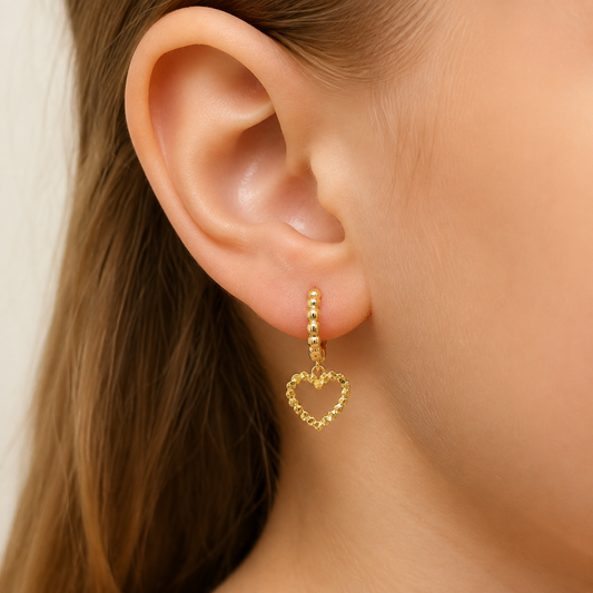 A close-up of a model's ear wearing a delicate gold huggie hoop earring with a dangling open heart charm.