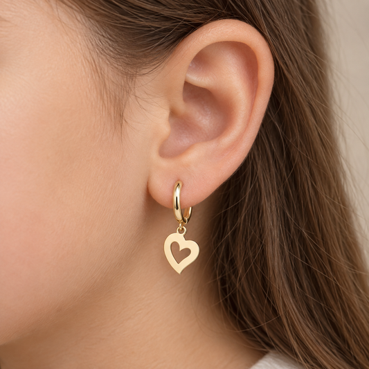 A close-up of a person's ear wearing a gold huggie hoop earring with a dangling open heart charm.