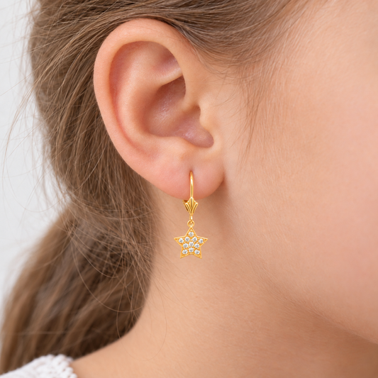 A close-up of a gold hoop earring with a dangling gold star charm embellished with small clear rhinestones, shown worn on a person's ear.