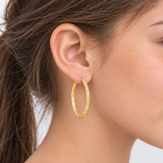 A close-up side profile of a woman wearing a textured gold hoop earring on her ear, with her hair tied back and a white top visible against a light gray background.
