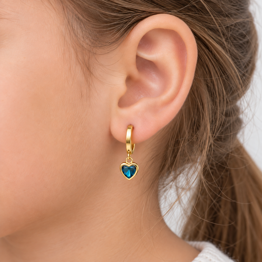 A close-up of a person's ear wearing a small gold huggie hoop earring with a dangling heart charm featuring a blue-green iridescent center.