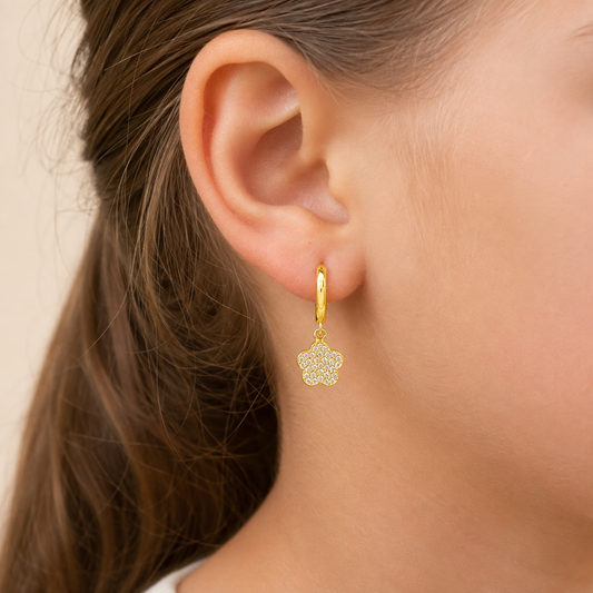 A close-up of a model's ear wearing a small gold huggie hoop earring with a dangling flower-shaped charm paved with clear stones.