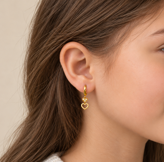 A close-up side profile of a woman wearing a delicate gold huggie hoop earring with a dangling open heart charm, set against a soft beige background.