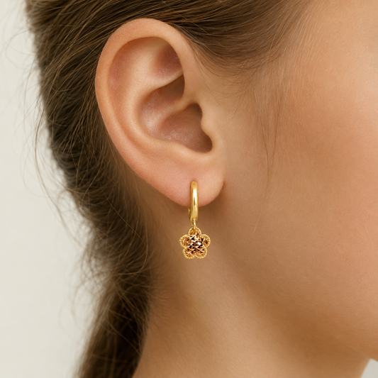 A close-up of a person's ear wearing a delicate gold huggie hoop earring with a small flower-shaped dangling charm set with tiny clear stones.