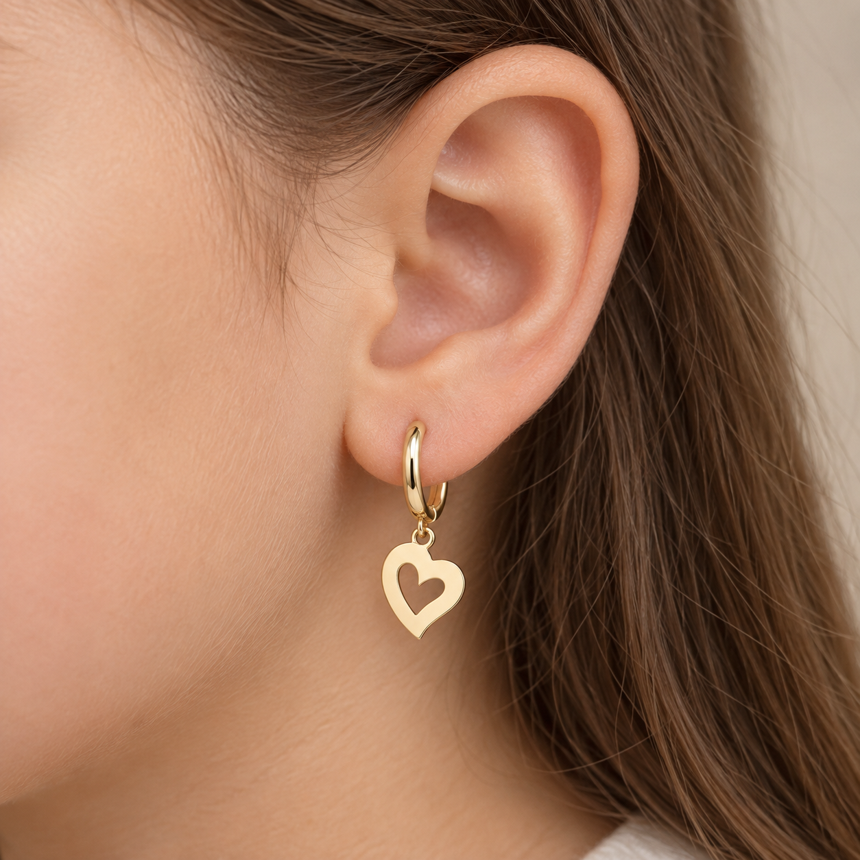 A close-up of a person's ear wearing a gold huggie hoop earring with a dangling open heart charm.