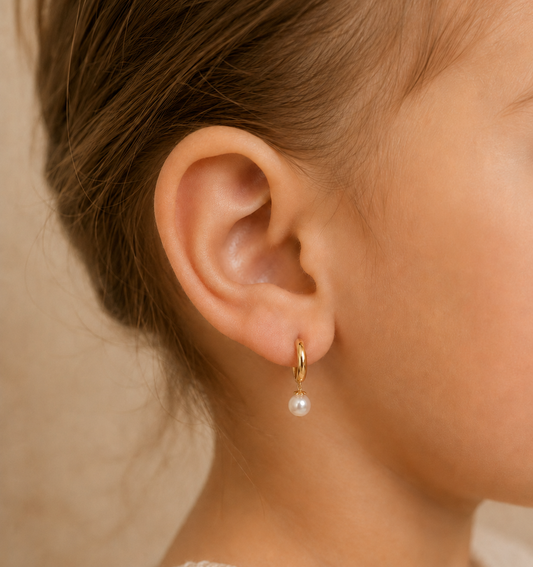 A close-up of a child's ear wearing a small gold huggie hoop earring with a dangling white pearl.