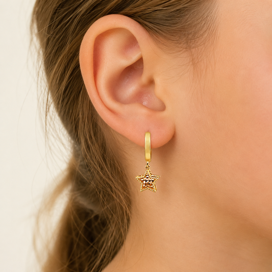 A close-up of a model's ear wearing a gold huggie hoop earring with a dangling textured star charm.