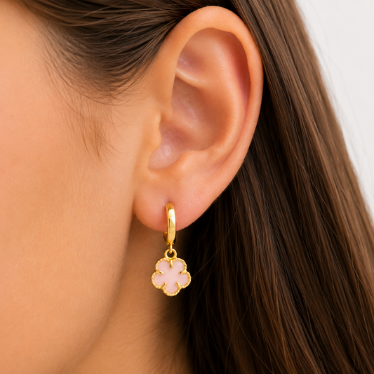 A close-up of a woman's ear wearing a delicate gold huggie hoop earring with a dangling four-petal flower charm in pale pink enamel.